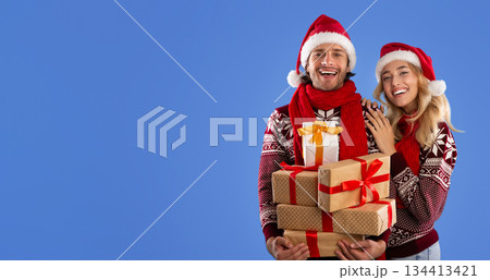 Portrait of adorable young couple in Christmas outfits holding heap of gift boxes over blue background Portrait of adorable young couple in Christmas outfits holding heap of gift boxes over blue background 134413421