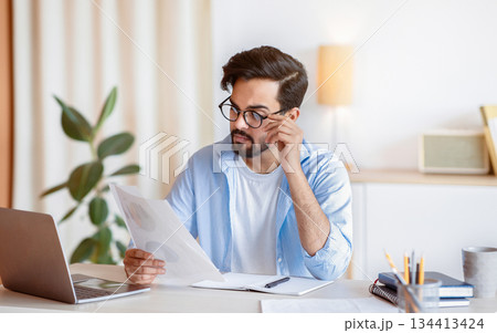 Young Eastern Businessman In Eyeglasses Working With Papers And Laptop In Office, Millennial Arab Entrepreneur Reading Reading Financial Documents While Sitting At Worplace, Free Space 134413424