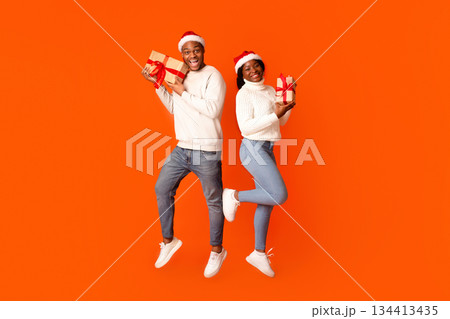 Cheerful Black Couple In Santa Hats Posing With Christmas Gifts, Holding Wrapped Present Boxes And Smiling At Camera While Jumping In Air Over Orange Studio Background, Full Length, Copy Space 134413435