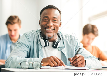 Modern Education Concept. Portrait of smiling African American male student sitting at desk in classroom at university, taking test or writing notes in his notebook, looking posing at camera Modern Education Concept. Portrait of smiling African American male student sitting at desk in classroom at university, taking test or writing notes in his notebook, looking posing at camera 134413456