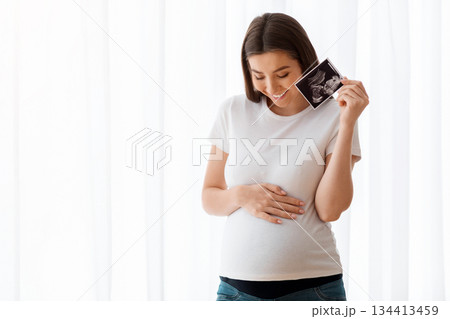 Happy Pregnant Woman Holding Ultrasound Sonography Scan Of Baby And Touching Belly While Standing Near Window At Home, Smiling Lady Enjoying Her Healthy Pregnancy And Maternity Time, Copy Space Happy Pregnant Woman Holding Ultrasound Sonography Scan Of Baby And Touching Belly While Standing Near Window At Home, Smiling Lady Enjoying Her Healthy Pregnancy And Maternity Time, Copy Space 134413459