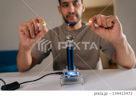Bearded student male demonstrating mini tesla coil generating wireless power, excitingly lighting up small light bulbs through electromagnetic induction, experimenting with electricity and physics. 134413472