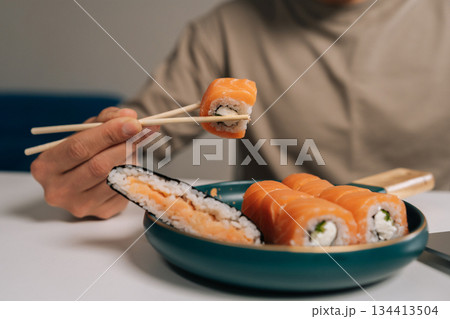Man using chopsticks to pick up fresh salmon and cream cheese sushi roll from plate of delicious Japanese maki, enjoying healthy and flavorful meal, closeup, unrecognizable person. Man using chopsticks to pick up fresh salmon and cream cheese sushi roll from plate of delicious Japanese maki, enjoying healthy and flavorful meal, closeup, unrecognizable person. 134413504
