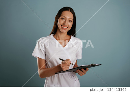 Notepad and pen in hands, nurse is standing. Young woman is in the studio against background Notepad and pen in hands, nurse is standing. Young woman is in the studio against background 134413563