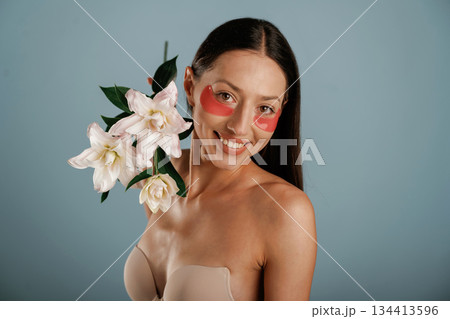 Cleaning patches under eyes, holding lily flowers. Young woman is in the studio against background Cleaning patches under eyes, holding lily flowers. Young woman is in the studio against background 134413596