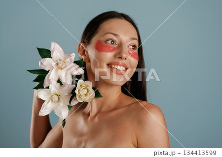 Cleaning patches under eyes, holding lily flowers. Young woman is in the studio against background 134413599