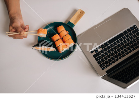 Top view of person enjoying takeaway sushi lunch with salmon and rice onigiri, using chopsticks from skillet plate positioned next to open laptop, representing modern remote work lifestyle. 134413627