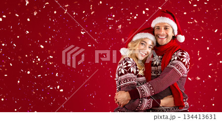 It's Christmas. Beautiful romantic couple in Santa hats and warm sweaters hugging on red background 134413631