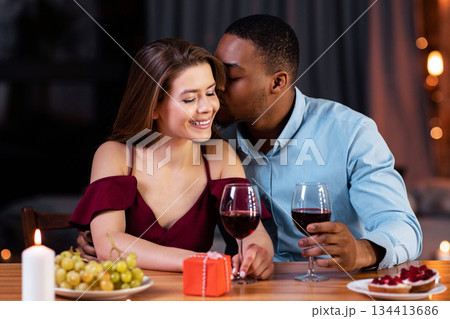 Multiracial couple in love dating and drinking wine during romantic dinner in restaurant, sitting at table, celebrating valentine's day together, black man kissing his happy girlfriend, copy space 134413686