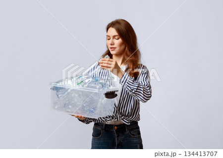 Young Beautiful Woman Putting Plastic Bottles To Container In Her Hands, Millennial Lady Collecting Waste For Recycling, Eco-Activist Sorting Garbage Over Light Studio Background, Copy Space 134413707