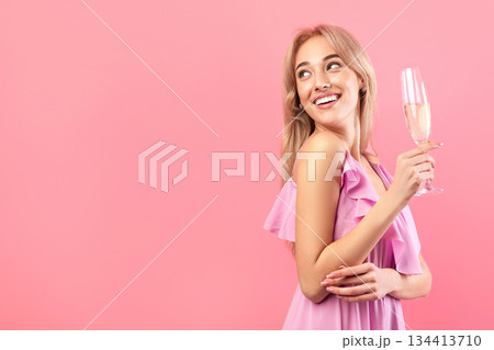 Beautiful blonde woman in dress holding glass of sparkly champagne, celebrating holiday, looking aside at empty space over pink studio background. Festive concept Beautiful blonde woman in dress holding glass of sparkly champagne, celebrating holiday, looking aside at empty space over pink studio background. Festive concept 134413710