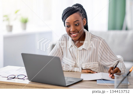 Young black lady having online training from home, using laptop and taking notes, copy space. Smiling african american woman attending online conference, writing tasks, living room interior 134413752