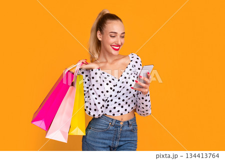 Looking And Searching For Discounts. Smiling female model carrying colorful shopping bags and using smartphone, making purchases in online store standing isolated on orange studio background Looking And Searching For Discounts. Smiling female model carrying colorful shopping bags and using smartphone, making purchases in online store standing isolated on orange studio background 134413764