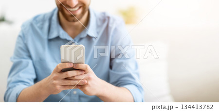 Happy Bearded Guy Using Smartphone Texting And Using Mobile Application On Phone Sitting On Couch At Home. Great App, Man Using Cellphone Browsing Internet Or Playing Game Indoor. Shallow Depth 134413782