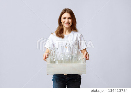 A young lady is smiling while holding a wooden crate filled with glass bottles for recycling. She promotes eco-friendliness with her involvement in sustainability activities. A young lady is smiling while holding a wooden crate filled with glass bottles for recycling. She promotes eco-friendliness with her involvement in sustainability activities. 134413784