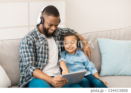 Black Father And Son Using Digital Tablet Computer Wearing Wireless Headphones Watching Movie Together Sitting On Sofa At Home. Weekend Leisure. Family, Gadgets And Modern Lifestyle Concept 134413808