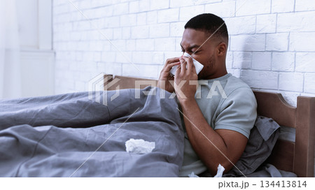 Sick African Man Blowing Runny Nose In Paper Tissue Lying In Bed At Home, Suffering From Rhinitis And Sinusitis. Ill Guy Sneezing Having Cold Symptoms Resting In Bedroom. Side View Sick African Man Blowing Runny Nose In Paper Tissue Lying In Bed At Home, Suffering From Rhinitis And Sinusitis. Ill Guy Sneezing Having Cold Symptoms Resting In Bedroom. Side View 134413814