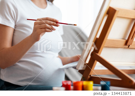 Pregnancy And Leisure. Unrecognizable Pregnant Woman Painting On Easel At Home, Expectant Mother Sitting On Couch And Drawing Picture, Enjoying Creative Pastime, Closeup Shot, Cropped 134413871