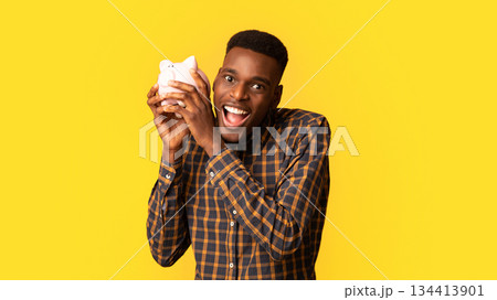 Funny african american guy shaking piggy bank, listening sound of coins, checking saved money, cheerful young black man enjoying economy, standing isolated over yellow background, copy space Funny african american guy shaking piggy bank, listening sound of coins, checking saved money, cheerful young black man enjoying economy, standing isolated over yellow background, copy space 134413901