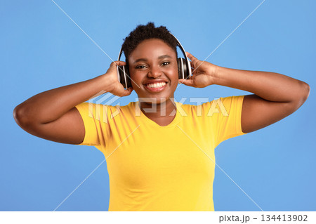 Music Lover. Happy Plus-Size African Woman Listening To Podcast Wearing Wireless Headphones Posing In Studio On Blue Background, Smiling To Camera. Great Playlist, Electronics Concept 134413902