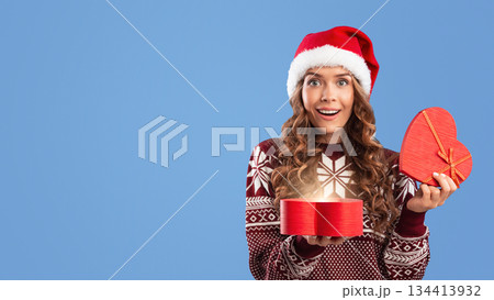 Christmas surprise. Gorgeous millennial woman in Santa hat opening heart shaped gift box on blue studio background, panorama. Young lady excited to receive her Xmas present Christmas surprise. Gorgeous millennial woman in Santa hat opening heart shaped gift box on blue studio background, panorama. Young lady excited to receive her Xmas present 134413932