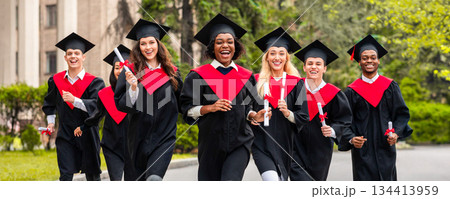 Cheerful multiracial students guys and young ladies in graduation costumes walking by university campus, holding diplomas and smiling. College, university graduation, education concept Cheerful multiracial students guys and young ladies in graduation costumes walking by university campus, holding diplomas and smiling. College, university graduation, education concept 134413959