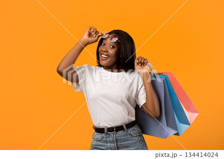 Stylish young black lady with sunglasses and shopping bags posing over orange studio background. Smiling african american woman removing glasses and holding purchases, copy space 134414024