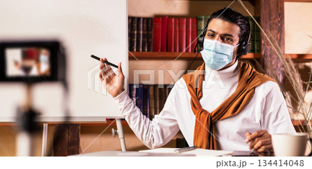 Distant Online Education. Indian Teacher Guy Making Video Lesson For Educational Blog Teaching English Grammar Rules, Pointing At Blackboard Sitting At Home, Wearing Face Mask. Selective Focus Distant Online Education. Indian Teacher Guy Making Video Lesson For Educational Blog Teaching English Grammar Rules, Pointing At Blackboard Sitting At Home, Wearing Face Mask. Selective Focus 134414048