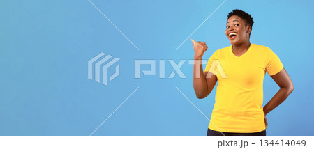 Look There. Cheerful Overweight Black Woman Pointing Thumb Aside At Copy Space Standing Posing On Blue Studio Background. African Female Points Finger Advertising Your Text. Panorama 134414049