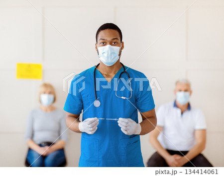 Portrait of black male doctor in protective mask and uniform holding syringe with coronavirus vaccine at hospital, patients waiting for covid-19 injection. Medical treatment concept Portrait of black male doctor in protective mask and uniform holding syringe with coronavirus vaccine at hospital, patients waiting for covid-19 injection. Medical treatment concept 134414061