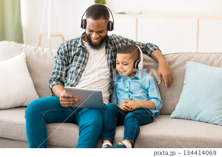 Young Black Father And Little Son Using Tablet Computer Together Wearing Wireless Headphones Watching Movie Sitting On Couch At Home. Family Of Two Browsing Internet Using Gadget Concept 134414069