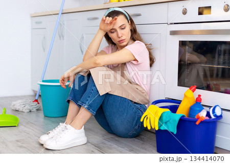 Depressed young housewife feeling tired from domestic chores, sitting on floor at kitchen. Young lady with cleaning supplies exhausted from house cleaning, suffering from OCD disorder 134414070