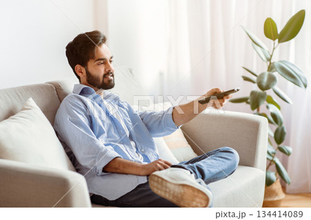 Arab man with remote controller switching channels while watching tv at home, young eastern guy relaxing on couch in comfortable living room, enjoying weekend pastime, copy space 134414089