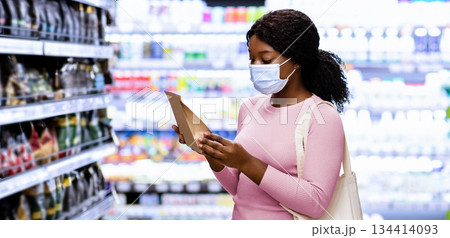 Millennial black lady with tote bag wearing face mask, shopping for groceries at mall. Young African American woman making choice of products, purchasing food at supermarket during covid outbreak Millennial black lady with tote bag wearing face mask, shopping for groceries at mall. Young African American woman making choice of products, purchasing food at supermarket during covid outbreak 134414093
