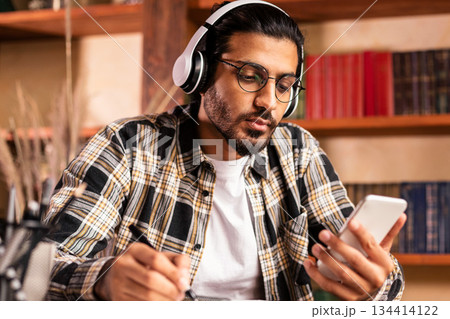 Indian Male Student Using Smartphone Learning Sitting At Books In University Library Indoor, Wearing Headphones. Educational Mobile App, Gadgets For College Study Concept. Selective Focus 134414122