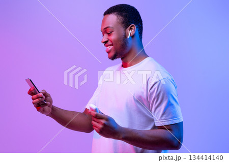 Tech Gadgets For Entertainment. Happy Black Guy Using Smartphone And Earpods For Listening Music, Holding Earphones Case In Hand, Cheerful African Man Standing In Neon Light Over Purple Background Tech Gadgets For Entertainment. Happy Black Guy Using Smartphone And Earpods For Listening Music, Holding Earphones Case In Hand, Cheerful African Man Standing In Neon Light Over Purple Background 134414140