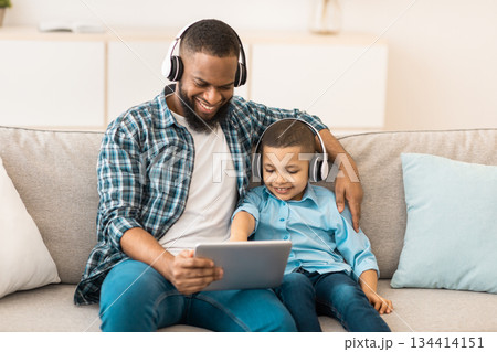 African Dad And Son Using Tablet Wearing Wireless Earphones Watching Movie Online Sitting On Couch Indoors. Family Using Computer Together Browsing Internet On Weekend. Gadgets Lifestyle African Dad And Son Using Tablet Wearing Wireless Earphones Watching Movie Online Sitting On Couch Indoors. Family Using Computer Together Browsing Internet On Weekend. Gadgets Lifestyle 134414151