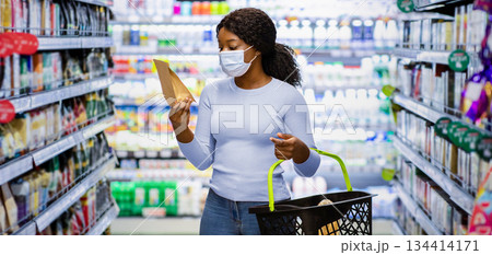 Young black woman in face mask shopping for groceries at huge supermarket. Millennial African American lady choosing products, buying food at mall during covid-19 lockdown Young black woman in face mask shopping for groceries at huge supermarket. Millennial African American lady choosing products, buying food at mall during covid-19 lockdown 134414171