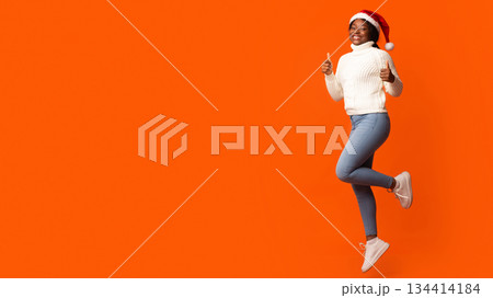 Holiday Offer. Joyful Black Santa Lady In Christmas Hat Jumping And Showing Thumb Up Gesture, Recommending Something, Having Fun On Orange Background In Studio, Cheering Xmas And New Year, Copy Space 134414184