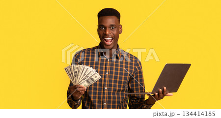 Online Lottery. Excited black guy with dollar cash and laptop celebrating success, overjoyed african american man holding money and computer, standing isolated over yellow background, free space 134414185