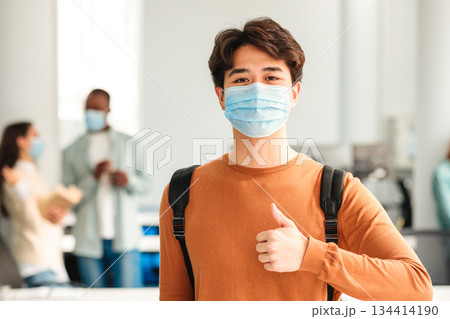 Protection Concept. Portrait of asian teenager wearing disposable surgical face mask to prevent infection, illness or covid-19, showing thumbs up sign gesture standing in classroom at high school 134414190