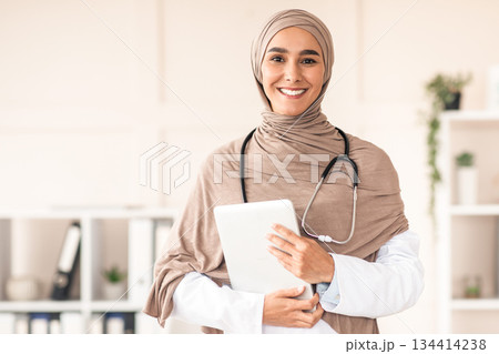 Modern technologies in healthcare concept. Happy female doctor in hijab posing with digital tablet in her office, copy space. Muslim woman therapist holding digital pad, clinic interior Modern technologies in healthcare concept. Happy female doctor in hijab posing with digital tablet in her office, copy space. Muslim woman therapist holding digital pad, clinic interior 134414238