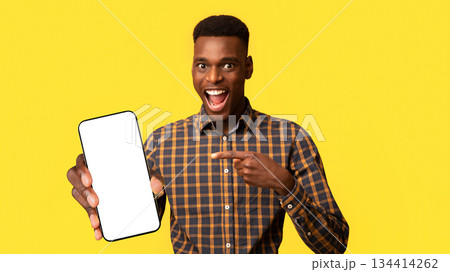 Mobile Offer. Excited Black Guy Pointing At Smartphone In His Hands, Emotionally Reacting To New App, Overjoyed Millennial African American Man Standing Isolated Over Yellow Background, Copy Space 134414262