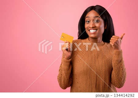 Happy black woman holding credit card and showing thumb up gesture on pink studio background. Beautiful African American lady recommending bank services. Electronic money concept Happy black woman holding credit card and showing thumb up gesture on pink studio background. Beautiful African American lady recommending bank services. Electronic money concept 134414292