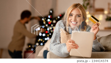 Pretty smiling woman shopping online on xmas, using credit card and digital tablet, sitting on sofa at living room. Positive lady buying presents online while her boyfriend decorating Christmas tree Pretty smiling woman shopping online on xmas, using credit card and digital tablet, sitting on sofa at living room. Positive lady buying presents online while her boyfriend decorating Christmas tree 134414374