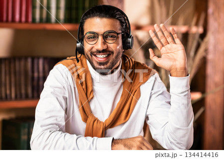 Happy Indian Man Wearing Headphones Waving Hello Smiling To Camera Sitting At Home. Hello, I'm Your Online Teacher. Modern Tutoring, Knowledge And Distance Education Concept 134414416