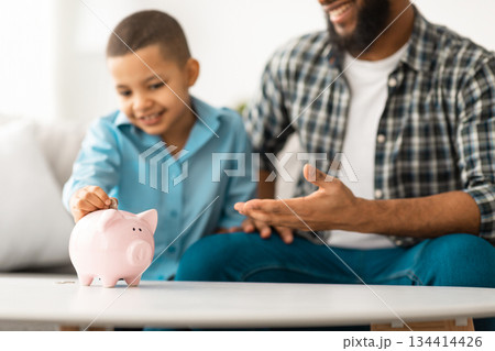 African Father And Little Son Putting Personal Savings Money In Piggybank Sitting Together On Sofa At Home. Selective Focus On Piggy Bank, Cropped. Dad Teaching His Kid Boy Financial Budget Planning African Father And Little Son Putting Personal Savings Money In Piggybank Sitting Together On Sofa At Home. Selective Focus On Piggy Bank, Cropped. Dad Teaching His Kid Boy Financial Budget Planning 134414426