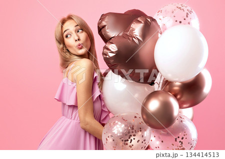 Beautiful blonde woman in cute dress holding balloons, pursing her lips for kiss, being playful on pink studio background, banner design with blank space. Party time, holiday celebration concept Beautiful blonde woman in cute dress holding balloons, pursing her lips for kiss, being playful on pink studio background, banner design with blank space. Party time, holiday celebration concept 134414513