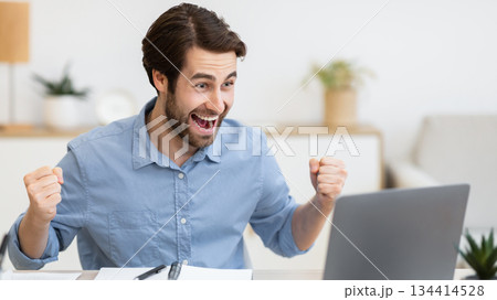 Excited Young Businessman At Laptop Computer Shaking Fists And Shouting Looking At Screen Sitting At Workplace In Modern Office, Celebrating Business Success. Victory Celebration, Successful Career 134414528