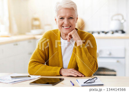 Happy senior woman freelancer working from home, sitting in kitchen, using digital tablet and smartphone, taking notes while reading financial report, smiling at camera, copy space 134414566